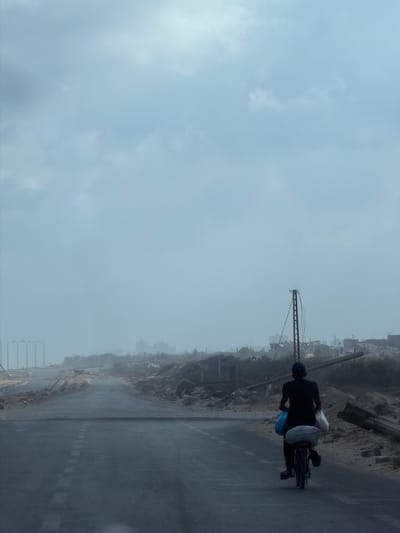 In Nuseirat, Palestinian Territories, a lone figure is seen from behind, riding a bicycle on a damaged road. The rider is wearing a dark t-shirt and pants, with a blue cap. They are carrying two large bags, one blue and one white, strapped to the back of the bicycle. The road itself appears cracked and partially covered in debris.

The surrounding environment is one of destruction. To the left, a row of streetlights stands against a hazy sky, with rubble and damaged structures in the middle ground. To the right, the landscape is similarly devastated, with piles of debris, broken wooden beams, and what appears to be the skeletal remains of buildings and infrastructure. A tall, metal structure, possibly a utility pole or part of a larger industrial framework, rises prominently on the right side of the scene. The overall atmosphere is somber and indicative of recent conflict or natural disaster. The sky is overcast with diffused light.
