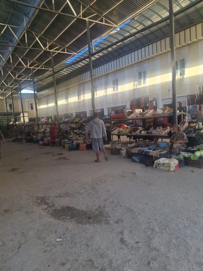 The image depicts an outdoor marketplace, likely in Ko`kdala, Uzbekistan, under a covered structure with a corrugated metal roof supported by metal beams and columns. Sunlight streams through gaps in the roof, casting long shadows across the scene. The ground is made of compacted dirt and concrete, with some wet patches and debris.
In the foreground, a man wearing a grey hoodie and blue shorts walks away from the camera. To the right, a woman in a patterned dress and headscarf is seated behind a table laden with produce. Various fruits and vegetables, including watermelons and cabbages, are displayed in crates and on the ground. Stalls with wooden shelves are visible, stocked with more goods. In the background, more market stalls and buildings with windows are present. Clothing and other items hang from racks. Other individuals can be seen browsing or working at the stalls, though they are less distinct. The overall impression is a busy, open-air market with a variety of goods for sale.