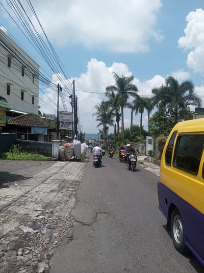 The image shows a street scene in what appears to be a city. There are several people on motorcycles driving down the road. There is a yellow and blue vehicle to the right. There is a building on the left of the image. There are several palm trees in the background. The sky is blue with white clouds. There are power lines stretching overhead. There is a sign for Villa Zodiac on the left side of the road. The road surface is cracked and uneven.