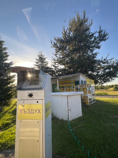 The image depicts an outdoor scene near a roadside structure in Dragushinovo, Bulgaria. In the foreground, a tall, light-colored machine with a yellow sticker bearing the Cyrillic word "ВЪЗДУХ" (meaning "AIR") is prominently featured. The machine has a reflective surface at the top, showing a sun glare. Adjacent to this machine, a coiled green hose lies on the grass, leading towards a white, shed-like structure. Behind the shed, a metal shelving unit is stocked with yellow gas cylinders. The shed itself has a sign with Cyrillic text, some of which appears to read " ЗА БИТОВИ ЦЕЛИ" (meaning "FOR DOMESTIC USE"). The background reveals a large evergreen tree, other trees, and a grassy field under a clear blue sky with a faint contrail. The overall impression is of a self-service air pump station or similar utility point, possibly associated with a small business or rest stop.