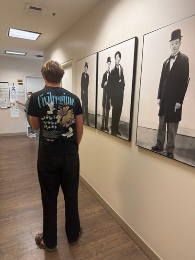 A young man stands in a hallway with his back to the camera, wearing a black t-shirt and dark jeans. The t-shirt features a graphic design with text and images of doves and flowers. He is also wearing sandals. To his left, a woman in a green shirt and a face mask stands near an eye chart on the wall. The hallway has wood-paneled flooring and neutral-colored walls. Several framed black and white photographs of what appear to be comedians in formal attire are displayed on the wall to the man's right. The setting appears to be a medical clinic or office. No specific location in Burbank, United States of America is identifiable from the image.