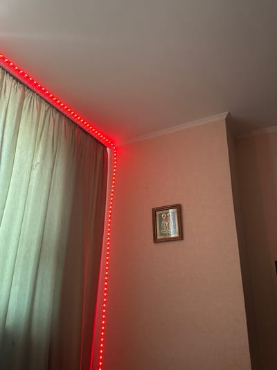 The image shows a corner of a room with a beige textured wall and a white ceiling. Red LED strip lights are installed along the top corner of the room, illuminating the ceiling and the wall. On the wall, there is a small framed icon of two religious figures, likely in an Orthodox Christian style. To the left, a light-colored curtain is visible, partially illuminated by the red lights. The scene appears to be an indoor domestic setting, potentially a bedroom or living area. No specific location in Criuleni, Moldova, can be determined from the visual information provided.