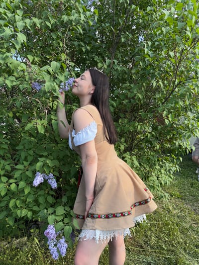 A woman in a tan dress with white lace trim and a floral ribbon design is smelling purple lilac flowers on a bush. The photo appears to be taken in an outdoor, grassy area. The city is Пальники, Russia.