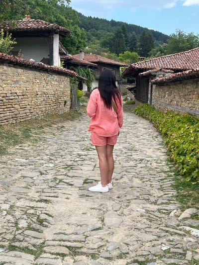 A woman stands on a cobblestone street in Zheravna, Bulgaria. She has long, dark hair and is wearing a coral-colored linen top and matching shorts, with white sneakers. She is facing away from the camera, looking down the street. The street is lined with traditional stone houses with red tile roofs. Lush green trees and vegetation are visible on the hills in the background. The sky is partly cloudy.