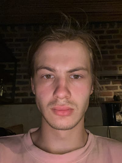 A young person with light brown hair and brown eyes is looking directly at the camera. They are wearing a pink t-shirt and have a light beard. The background appears to be an indoor setting with a brick wall behind them. The lighting is dim, creating shadows on their face. The scene is not in Nocaima, Colombia.