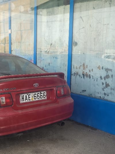 The image displays the rear of a stationary red Toyota Celica automobile parked on a dark gray asphalt surface adjacent to a building structure in Ανδραβίδα, Greece. The vehicle is positioned with its rear angled towards the viewer, featuring a pronounced spoiler and a single exhaust pipe extending from the lower right of the bumper. A Greek license plate, centrally mounted, displays the alphanumeric sequence "HAE-6666". The adjacent building wall is composed of multiple vertical, translucent panels, likely glass or plastic, separated by blue vertical framing elements. These panels exhibit significant soiling, with visible streaks, smudges, and accumulated grime. Below the translucent panels, the lower portion of the wall is a solid blue color. Faint reflections of an external environment, possibly trees or distant structures, are visible on the upper sections of the panels. No individuals are present in the scene.