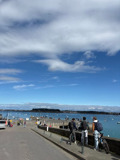 The photo was taken at 48.631, -2.052, which is the location of the port in Dinard, France. The image shows a paved road along the water with a group of people and bicycles. Two people are riding bicycles and the others are standing on the side of the road. The day is sunny with a clear blue sky and white fluffy clouds. The water is calm and there are sailboats on the water. The scene appears to be on the edge of a town or city, with a landmass in the distance.  There is a stone wall on the right side of the photo separating the road from the water. There is a car on the left side of the road. The road leads down to the pier where several people are walking.
