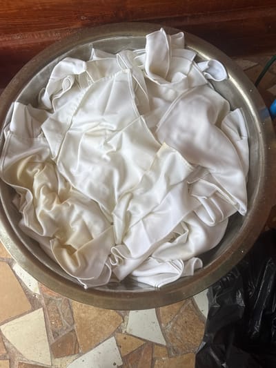 An overhead view reveals a large, circular, metallic basin filled with numerous pieces of white and off-white fabric. The fabric is wet, crumpled, and exhibits localized yellowish-brown discoloration. The basin's interior and rim show signs of wear. The basin rests upon a floor composed of irregularly shaped tiles in shades of brown, beige, and light gray. Behind the basin, in the upper portion of the image, a reddish-brown wooden surface with prominent grain is visible. To the right of the basin, a crumpled black plastic bag is present on the tiled floor. A small segment of a blue object, possibly a hose, is visible in the upper right corner. This scene is located in Tela, Honduras.