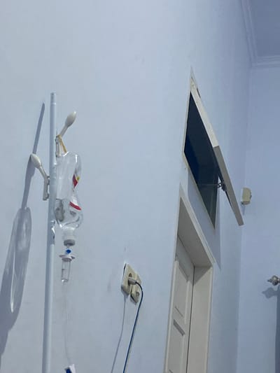 The image depicts an indoor scene, likely a hospital room, in Walennae, Indonesia. Dominating the left side of the frame is a medical IV stand. An IV bag, partially filled with clear fluid, is suspended from the stand, connected to tubing that leads downwards out of view. The bag has red and white labeling. To the right of the IV stand, a wall-mounted electrical outlet with a plug and blue and white wires is visible. Further to the right, a portion of a doorway with a white door is seen, and above it, a small, open window is angled upwards. The walls are painted a light blue. The lighting suggests artificial illumination, casting shadows from the IV stand and other objects.