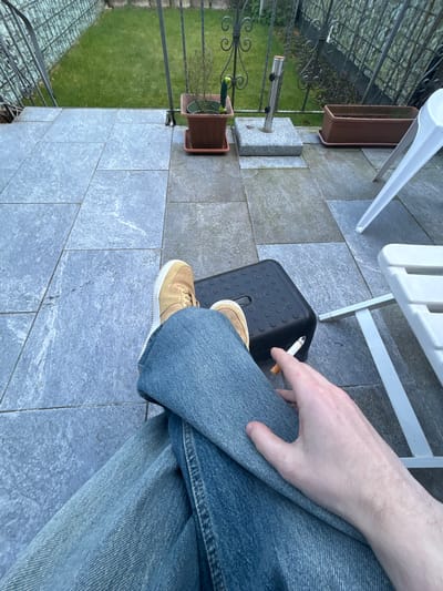 The image depicts an outdoor patio scene in Hagen, Germany, viewed from a first-person perspective, looking downward. In the foreground, the lower body of an individual is visible, clad in light blue denim jeans. Their legs are crossed, with the right leg over the left. The individual's feet, wearing tan-colored sneakers with white soles, are propped on a black plastic step stool with a textured top surface. The left hand of the individual rests on their left thigh, holding a lit cigarette between the index and middle fingers.

To the right of the individual, a portion of a white plastic chair with a horizontal slat is visible. The patio floor is constructed from large, rectangular grey stone tiles, some appearing lighter and others darker or damp. In the midground, a dark metal fence with decorative scrollwork at the base and a grid pattern above separates the patio from a grassy lawn area. Behind the fence, a brown potted plant with thin branches is situated on a terracotta-colored saucer, with a green gardening tool inserted into the soil. Adjacent to this plant is a rectangular grey granite base for an umbrella stand, featuring a silver-colored metal pole. Further to the right, an empty, long, brown rectangular planter box with a saucer is visible.