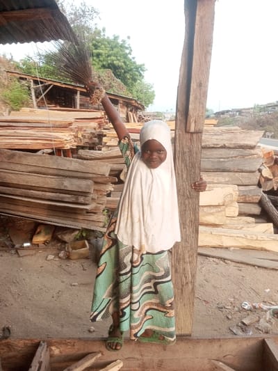 A young girl wearing a light-colored hijab and patterned dress stands amidst stacks of lumber. She holds a bundle of sticks above her head with one arm raised, while the other hand rests on a wooden post. The setting appears to be a woodyard or construction site, with a partly visible structure behind the wood stacks and greenery in the distance. The scene is captured in daylight.