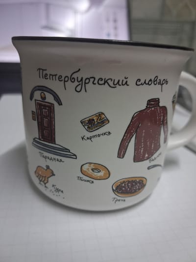 The image shows a close-up, slightly angled view of a white ceramic mug. The mug is decorated with various hand-drawn images and Russian text. The text at the top reads "Петербургский словарь" which translates to "Petersburg Dictionary". Below this, there are several illustrations: a door with a doorbell and steps labeled "Парадная" (Entrance), a postcard labeled "Карточка" (Card), a brown turtleneck sweater labeled "Бадлон" (Sweater), a chicken labeled "Кура" (Chicken), a bagel labeled "Пышка" (Doughnut/Bun), and a dish of food labeled "Греча" (Buckwheat). The mug has a dark rim and a handle is visible on the right side. The background is blurred and appears to be an indoor setting with some electronic equipment visible, possibly a computer monitor. There is no indication that this image is related to the city of Zhodzina, Belarus.