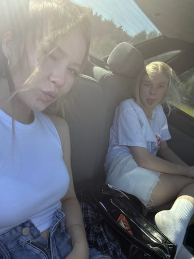 Two young blonde women are seated in the back of a car, with one woman in the foreground and the other behind her. The woman in the foreground is lying down with her head resting on the car seat, looking at the camera with her lips pursed. Her hair is blowing slightly, and she is wearing a white tank top and denim shorts. The woman behind her is also lying down and sticking her tongue out at the camera. She is wearing a white t-shirt and white shorts. The setting appears to be daytime, with sunlight streaming into the car. The windows show a blurry background of trees and sky, suggesting a rural or suburban location. The mood appears to be casual and playful, with the women making silly faces for the photo. There is a black bag with a zipper and some items inside visible between the two women, and a plaid shirt also partially visible.