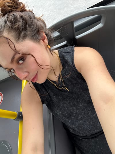 The image captures a young woman from a sideways perspective, appearing to be lying down or leaning inside what looks like a public transport vehicle, possibly in Apodaca, Mexico. She is the central subject, looking directly at the camera with a neutral expression.
She has medium-brown, curly hair, pulled back into a bun with several loose, damp-looking strands framing her face. She wears a dark grey or charcoal-colored sleeveless top. Around her neck are multiple thin golden necklaces, one of which features a small cross pendant. She also has a golden ear cuff visible on her left ear. Her skin tone is light.
The setting consists of grey plastic seating and a bright yellow metal handrail, common features in buses or trains. Below the handrail, a dark surface shows a partially visible white circular sticker with red, unreadable text. The lighting appears to be bright and even, suggesting daytime conditions. Some of her curly hair strands are visible on the grey surface of the seat near her arm. No specific weather conditions are discernible, but the indoor setting implies protection from the elements.