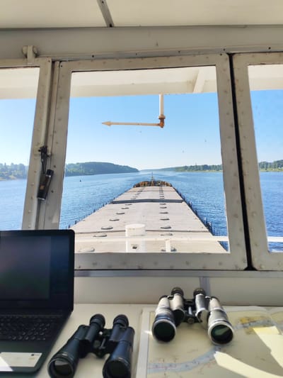 The image is taken from the bridge of a barge, looking out through a large window. The barge is traveling on a wide, blue river with tree-lined shores. The sky is clear and blue. On the deck of the barge, there are several circular hatches visible. In the foreground, on a white surface, there is a black laptop and a pair of black binoculars placed on a nautical chart. There is also another pair of binoculars visible on the white surface. A white directional arrow is mounted on a pole outside the window, pointing to the left. The scene depicts a clear, sunny day on a waterway, likely the river near the city of Pesochne, Russia, given the context of the image location.