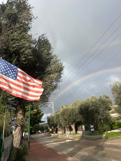 The image captures a street scene in Beit Yitshak Sha'ar Hefer, Israel, under a cloudy sky with visible atmospheric phenomena. In the foreground, a large American flag, displaying red and white stripes and a blue canton with white stars, is unfurled from a flagpole on the left side, partially obscured by the dense, silvery-green foliage of a mature olive tree. Below the American flag, several smaller Israeli flags are visible on a lower pole or fence. The street, paved with asphalt, recedes into the distance, bordered by a reddish-brown brick sidewalk on the left and a row of large, gnarled olive trees planted in raised stone planters on the right. Utility poles and overhead power lines traverse the sky above the street. In the mid-ground and background, a distinct double rainbow arches across the grey sky, with the primary bow clearly defined and a fainter secondary bow visible above it. No individuals are present in the scene.