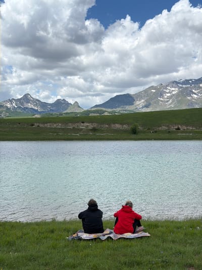 This image depicts a serene outdoor scene in the city of Javorje, Montenegro, featuring two individuals observing a tranquil lake against a backdrop of distant mountains.

In the foreground, two individuals are seated from behind on a patterned blanket spread on a vibrant green grassy bank that slopes gently towards the water. The person on the left wears a dark, possibly black, hooded sweatshirt, while the person on the right wears a bright red hooded sweatshirt. Both are facing away from the viewer, looking towards the lake and the landscape beyond.

The midground is dominated by a wide, calm body of water, likely a lake, exhibiting a light blue-green hue with subtle ripples across its surface, suggesting a gentle breeze. Beyond the lake, a strip of undulating green land rises, forming a gentle hill or embankment.

The background features a majestic mountain range under a partly cloudy sky. The mountains are rugged, with visible rock formations and patches of snow, indicating a significant elevation. Some mountains have tree lines at their bases, appearing as dark green patches. The sky above is a mix of bright white and grey cumulus clouds, with intermittent patches of clear blue sky visible, particularly in the upper right quadrant. The overall lighting suggests a daylight scene, possibly overcast but with some ambient brightness.
