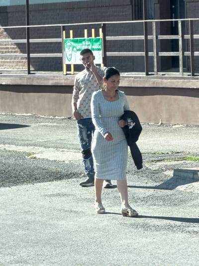 The image captures a man and a woman standing on a paved surface adjacent to a building in Liptovský Hrádok, Slovakia. The building is multi-story with a distinctive window design. The man is wearing a patterned shirt and distressed denim shorts. The woman is wearing a light-colored, knee-length dress and light-colored shoes. A black item, possibly a bag or hat, is placed on the woman's back. A green banner with white lettering (containing the words "VI LIGA" and "SRN LUPTOVSKY HRADOK") is attached to the building. The location appears to be an urban environment with sunlight casting shadows.