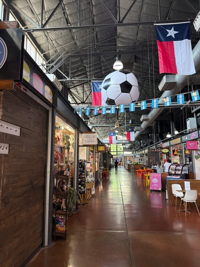The image depicts an indoor marketplace with a high ceiling supported by industrial metal beams. The ceiling is insulated with dark material. Fluorescent lights are visible, as are various pipes and ventilation systems. The space is decorated with flags, including a Texas flag and several small Argentinian flags strung across the ceiling. A large inflatable soccer ball is suspended from the ceiling.
The marketplace floor is a polished, reddish-brown concrete with visible seams. The sides of the walkway are lined with various stalls or shops. On the left, there is a stall with wooden paneling and a dark sign. Further down, a stall displays merchandise on shelves, including what appears to be clothing or accessories, decorative items, and bags. A sign above this stall reads "Folklore & Tradition". On the right, there are food stalls with display cases, tables, and chairs. One stall has a pink sign that says "BOOST YOUR ENERGY The 'Chocolat Rainbow Bowl'".
Several people are present in the scene, walking or browsing. A person in a dark shirt and pants walks away from the camera down the central aisle. In the distance, other individuals are seen near the food stalls. The overall atmosphere suggests a bustling, indoor market environment with a mix of retail and food vendors, possibly in a historic or repurposed industrial building. The presence of the Texas flag and Argentinian flags suggests a possible theme or connection to these regions, perhaps related to soccer given the prominent soccer ball. Without additional context, pinpointing the exact location in Dallas is not possible based solely on the visual information.