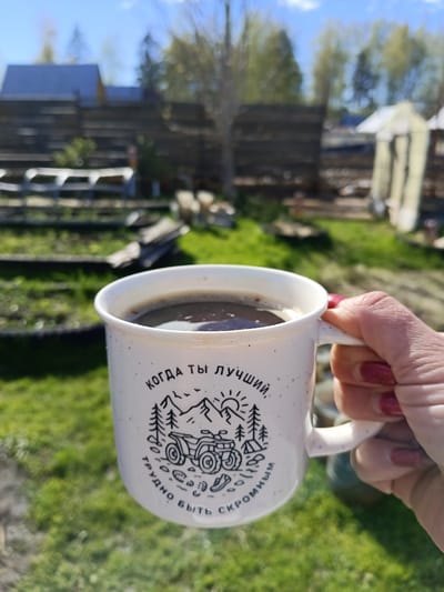 A woman's hand with red nail polish holds a white mug filled with dark liquid, likely coffee, in front of a blurred outdoor scene. The mug has a design featuring a mountain, trees, and an ATV. In the background, there's a wooden fence, a greenhouse-like structure, and green grass. The location is Вичелово, Russia.