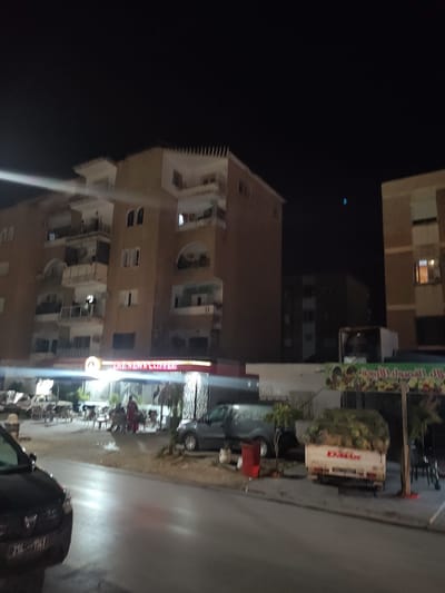 At night, a street scene in Ariana, Tunisia, shows a multi-story building with balconies and arched windows. A lit sign above a storefront reads "CHEZ NEWN COFFEE". In front of the building, outdoor seating with tables and chairs is visible, with a few people seated. To the right, a small white truck filled with produce is parked. A dark car is in the foreground on the left, with its license plate partially visible. Another vehicle, a gray SUV, is parked to the right of the truck. The street is paved, and there are trees and planters along the sidewalk. The sky is dark and appears clear.