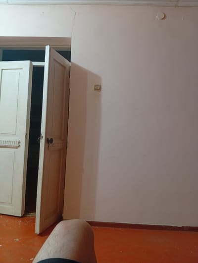 The image depicts an interior scene, likely a residential room, in Жартытөбе ауылдық округі, Kazakhstan. The room features a light-colored wall with a visible crack near the ceiling on the right side, a white crown molding along the top edge, and a dark brown baseboard along the bottom. An open doorway with two white wooden doors is prominent on the left side of the frame. The doors have decorative molding and dark circular doorknobs. The floor is a rust-orange color with scattered white debris. In the foreground, a portion of a person's leg and knee is visible, with the leg angled towards the left. The lighting is dim, casting soft shadows. A light switch or outlet is present on the wall to the right of the doorway.