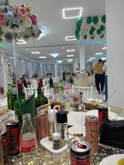 The image captures a festively decorated event space, possibly a wedding hall, filled with people. Long tables stretch into the background, adorned with white tablecloths and chairs. The ceiling is illuminated by modern, rectangular light fixtures and decorative crystal chandeliers. Green and white balloons are attached to the walls, adding to the celebratory atmosphere. The focus of the image is a table laden with various items: bottles of alcohol including Jameson whiskey, cans of Xixo ice tea and Hell energy drinks, and condiments such as salt and pepper shakers. The table is topped with a sparkly gold tablecloth, suggesting a luxurious setting. People are visible in the background, both standing and seated at the tables. Based on the OCR, there is a bread company name (Хлебозав Лозница) and a Bulgarian word (ПИРИН). The scene appears to be indoors at night, with a crowd celebrating a festive event. There is no evidence indicating this event or location is in Bulgaria.