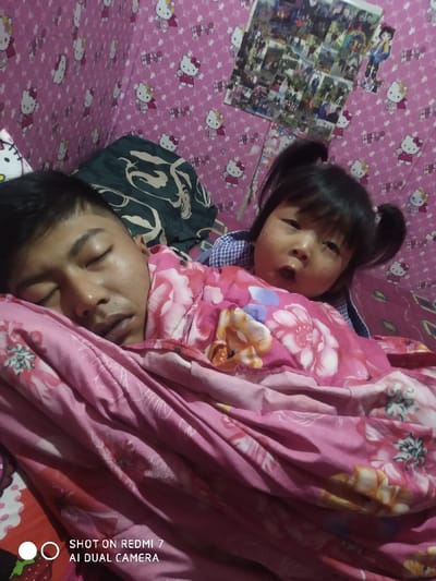 A man is asleep under a pink floral blanket. A young child with pigtails sits behind him, looking directly at the camera. The room's wallpaper features a Hello Kitty design, and a collage of photos is visible on the wall above them. The scene appears to be indoors, possibly a bedroom. The image includes a watermark at the bottom, indicating it was taken with a Redmi 7 AI Dual Camera.