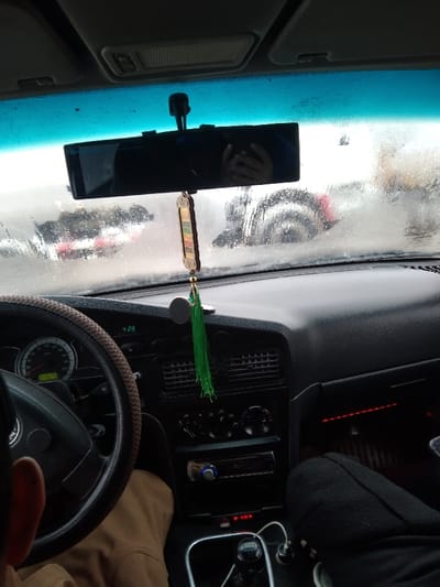 The image captures the interior of a vehicle in Karaulbazar, Uzbekistan, looking forward through a wet windshield that obscures the external environment due to precipitation or heavy condensation. Two individuals are present in the front cabin. The driver, partially visible on the lower left, shows an ear, shoulder, and light brown pants. On the lower right, the front passenger's lower leg, clad in black pants and a dark shoe, is visible. A reflection of hands holding a camera or smartphone is seen in the centrally mounted rearview mirror, indicating the photographer. The dark dashboard features a steering wheel with a brown patterned cover on the driver's side. The instrument cluster behind the steering wheel displays an odometer reading of "359203" and a digital time of "4:28". The center console includes air vents, rotary climate controls, an "AUTOSOUND" brand stereo unit, and a manual gear shifter. Multiple wires are connected to auxiliary power outlets near the gear shifter. A decorative ornament, consisting of a rectangular, multi-colored wooden piece and green tassels, hangs from the rearview mirror. A circular magnetic mount is affixed to the dashboard to the right of the ornament. Red ambient lighting is visible within the passenger footwell. Through the wet and obscured windshield, blurred outlines of other vehicles, including a red component and a large wheel, are faintly discernible in the background.