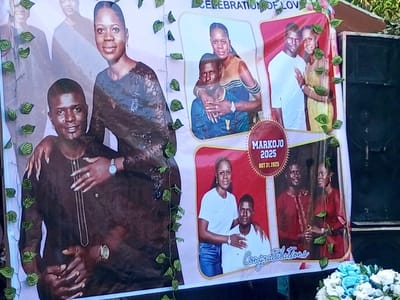 A large commemorative banner featuring an African couple is displayed outdoors in Amokvo, Nigeria. The banner is adorned with green artificial leafy vines and showcases multiple photographs. The primary image on the left depicts the woman, wearing a dark patterned top and a watch, standing behind the man in a dark brown traditional garment, with her arm around his shoulder and hand on his chest. Both are smiling. Other images on the right side of the banner show the couple in various poses and attire: embracing, standing side-by-side, and the woman in casual wear next to the man in a polo shirt. Text on the banner includes "CELEBRATION OF LOVE," a red box with "MARKOJO," "2025," and "OCT 31 2025," and "Congratulations" in cursive. To the right of the banner, a dark, large speaker is visible, and at the bottom right, a small arrangement of light blue and white artificial flowers is present.