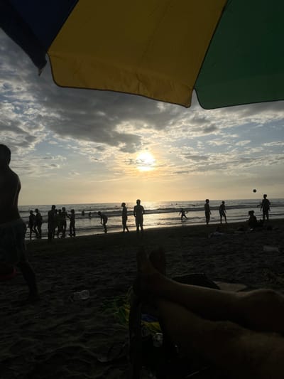 The scene takes place on a beach in Montanita, Ecuador, located at -1.824 latitude and -80.755 longitude. The image is captured from behind a beach umbrella, the bottom of which is visible in the foreground. The foreground features the legs of a person sitting in a chair, with a dark shape of another chair and a bright yellow beach towel behind them. In the distance, a group of people are silhouetted on the beach against a bright, cloudy sunset. Some of these people are standing, and some appear to be walking or playing a game of beach volleyball, as a ball is seen in the air. The beach is sandy, and the ocean is visible in the background. The overall tone of the image is serene and peaceful.