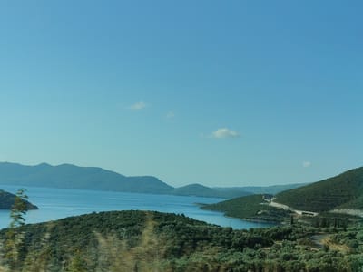 This image depicts a scenic view of the Municipal Unit of Pelasgia, Greece, likely taken from an elevated vantage point. The foreground is dominated by lush, green vegetation, characterized by dense trees and shrubs, with some appearing slightly blurred, suggesting movement such as a vehicle passing by. This foliage covers rolling hills that slope down towards a body of water.
In the middle ground, a calm, blue sea or large lake is visible, with several verdant islands or peninsulas jutting out into it. The coastline is irregular, creating coves and inlets. In the distance, a range of larger, mountainous islands or landmasses form a hazy backdrop against the horizon. The sky is a clear, pale blue with a few wispy white clouds scattered across it.
A paved road with guardrails is visible winding its way up the side of one of the hills in the right mid-ground. There are also some structures, possibly buildings or infrastructure related to the road, visible on the hillside. The overall impression is one of a serene, natural landscape with a touch of human development in the form of the road. There are no people visible in the image.