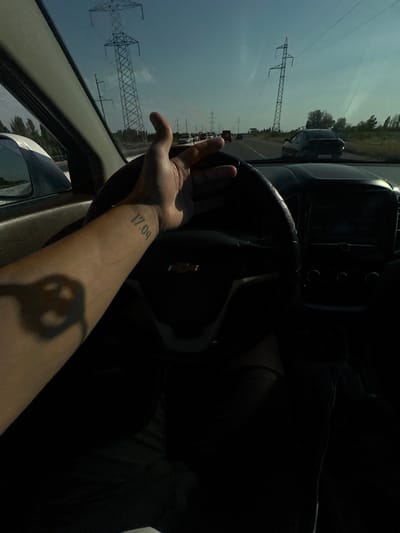 A first-person perspective from inside a car shows a driver's left arm and hand resting on the steering wheel. The arm has a tattoo of "17.09" on the forearm. The steering wheel is black and appears to be from a Chevrolet. The car's dashboard and a portion of the infotainment screen are visible. Outside the car, through the windshield, a clear sky with some clouds is visible. Several tall electrical transmission towers are prominent in the background, along with trees and other cars on a highway. The scene appears to be a daytime shot, possibly during the day. The location is identified as Istiqlol, Uzbekistan.