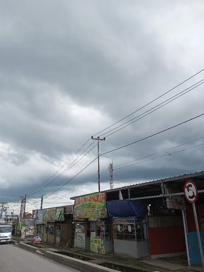 This image displays a roadside commercial area in Cibadak, Indonesia, under an overcast sky filled with dense grey clouds. The foreground features a paved road with a concrete curb and a narrow drainage channel alongside it. A white minivan is partially visible on the left side of the road. To the right, a row of semi-permanent commercial stalls is present, constructed with corrugated metal roofs and some blue tarpaulin coverings. These stalls exhibit multiple brightly colored advertising banners, notably one green banner proclaiming "BAKSO SOTO MURAH." A red and white circular road sign, likely indicating "No Stopping" or "No Parking" (a red 'S' with a diagonal line), is affixed to a light blue pole on the far right. Numerous utility poles support a complex network of overhead power lines that crisscross the frame. In the middle-ground, a reddish-white telecommunications tower stands prominent behind the stalls. No individuals or specific activities are clearly discernible within the scene.