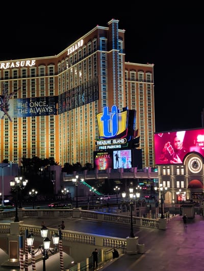 An eye-level, night time, long shot captures the exterior of the Treasure Island Hotel and Casino on the Las Vegas Strip in Paradise, Nevada, United States of America. The hotel, a large, orange, multi-story building with white shutters on its windows, is illuminated by the surrounding city lights. A large, blue and yellow sign with "TI" is prominently displayed on the front of the building, alongside other illuminated signs advertising "Treasure Island" and "Free Parking". To the right of the main hotel, a large, bright pink digital billboard displays an image of a magician holding playing cards. Below this billboard, there are smaller illuminated signs for Cirque du Soleil's "Mystere" and other attractions. A bridge with decorative lampposts spans a body of water in the foreground, with several people visible on the bridge and surrounding walkways. Cars are seen driving on a street behind the bridge. The scene is set against a dark night sky.