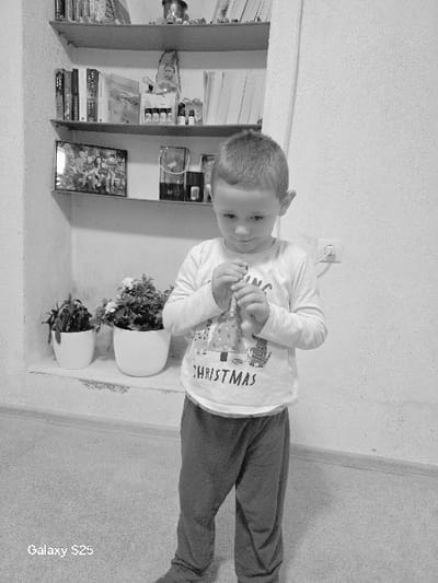 An indoor, black and white image depicts a male child standing in a room in Karnalovo, Bulgaria. The child, dressed in a long-sleeved shirt featuring a 'CHRISTMAS' graphic and dark trousers, stands facing forward with his gaze directed downwards at a slender, light-colored object he manipulates with both hands at chest level. To the child's left and rear is a recessed wall niche containing three shelves. The shelves are stocked with various books, a framed photograph of multiple individuals, small figurines, and an assortment of bottles and containers. On a lower ledge within the niche, two white potted plants are positioned. The adjacent wall to the right of the child displays a visible two-pin electrical outlet. The floor surface appears light-colored and textured.