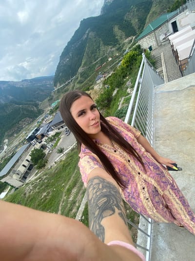 A woman with long, dark hair is taking a selfie on a concrete balcony overlooking a village nestled in a steep, mountainous valley. The village appears to be Chalda, Russia, characterized by its buildings with dark, sloped roofs and roads winding along the terrain. The woman is wearing a colorful, patterned dress and has a tattoo on her left arm. She is holding a phone in her right hand, and her left hand rests on a metal railing. The sky is overcast with clouds. The buildings in the village range from multi-story structures to smaller houses, with greenery and trees interspersed throughout the landscape. The overall scene depicts a scenic, remote location with a focus on the woman and the expansive mountain views.