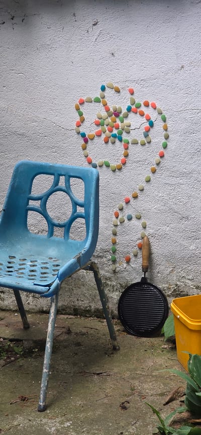 An outdoor scene in Partizanin, Bulgaria, displays a weathered blue plastic chair, a pattern of colorful stones on a white wall, and various utilitarian objects on concrete ground.

A dilapidated light blue plastic chair with a metal frame occupies the left foreground, its seat and backrest exhibiting significant wear, fading, and chipping. The plastic backrest features a decorative pattern of interconnected circular and arched cut-outs, while the seat is perforated with small rectangular holes. The metal legs, painted a faded light green-blue, show extensive paint loss and rust.

On the white, textured wall behind the chair, a mosaic of small, oval-shaped stones in various colors (yellow, green, orange, blue, pink, purple, white) is adhered. These stones form a central floral or star-like pattern, from which a curving trail of single stones extends downwards and to the right. The wall itself shows cracks and areas of discoloration, indicative of age and exposure.

Below the stone pattern, resting on the concrete ground against the wall, is a black circular cast-iron griddle pan with a ridged surface and a short, light brown wooden handle. To its right, partially visible, is a bright yellow plastic bucket or container. The concrete ground is uneven, marked with cracks, dirt, and sparse organic debris including small leaves and pine needles. Green foliage is present at the far right and bottom edge of the frame.