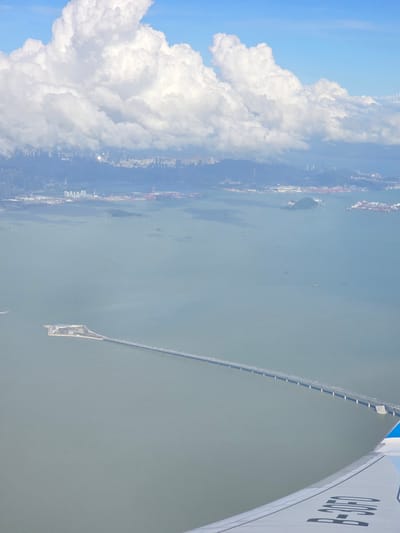 An aerial view captures the vast expanse of the Pearl River Delta, with the HZMB bridge stretching across the calm, grayish-green water. The bridge, a prominent engineering feat, connects to an artificial island which is surrounded by a breakwater. In the distance, a sprawling cityscape with numerous buildings is visible under a bright blue sky dotted with large, fluffy white clouds. Several ships and smaller vessels are scattered across the water. The wing of an airplane is visible in the foreground, with partial registration markings indicating "B-3050".