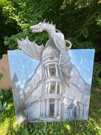 This media file shows a painting on a flat surface, possibly a canvas or wood panel, depicting a dragon in front of a building that resembles Gringotts Bank. The dragon is silver and detailed with wings, claws, and a long neck. The building behind it is painted in shades of gray, with columns and arched windows. The words "GRINGOTTS BANK" are visible on the building. The background is painted with a light blue sky.
The painting is set outdoors, with lush green foliage and grass surrounding it. The lighting suggests it is daytime, with sunlight filtering through the leaves. The scene appears to be in a garden or yard. There are no people visible in the image. The overall impression is that of a creative piece displayed outdoors. The painting is positioned at an angle, making it difficult to discern any specific activity or event. The setting does not provide strong cues to the exact location beyond being outdoors.