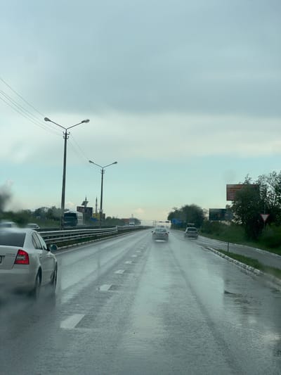 The image shows a highway scene in the city of Чистенское сельское поселение, Ukraine, during a rainy day. The road surface is wet and reflective, indicating recent or ongoing rainfall. Multiple vehicles are visible on the road, including cars and a van, moving in the same direction. Road markings are present and appear as white dashes. Lampposts are visible on the side of the road, and some signage can be seen alongside the highway. The sky is overcast, suggesting a cloudy or stormy weather. The overall impression is one of driving on a wet highway under gray skies.