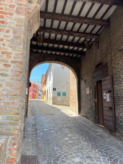 The image depicts an arched stone passage in the town of Barbara, Italy, opening onto a narrow cobblestone street lined with colorful buildings. The passage is covered by a wooden beamed ceiling with two antique lanterns hanging from it. The walls of the passage are constructed of rough-hewn brick. On the right wall, there is a large, dark wooden door, partially open, revealing a dark interior. A sign is posted to the right of the door. Further down the street, buildings in shades of yellow, pink, and white are visible under a clear blue sky. The cobblestone street shows some signs of wear and tear, with a few manhole covers visible. The overall impression is of a historic Italian town street on a sunny day.
