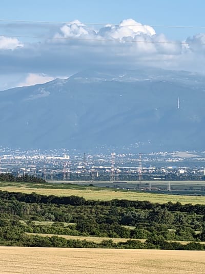 This image depicts a panoramic view of a city and a mountain range under a sky filled with clouds. In the foreground, there are fields of dry, golden crops. Behind the fields, a dense line of green trees forms a natural barrier. Further in the distance, industrial structures with red and white coloring are visible, along with a city skyline characterized by numerous buildings and what appears to be a tall communication tower on the mountain in the background. The mountain range itself is vast and hazy, with patches of lighter terrain, suggesting natural features or snow. The overall scene captures a landscape where nature and urban development meet, with a prominent mountain serving as a backdrop to the city and industrial complex. The location is Bogyovtsi, Bulgaria.