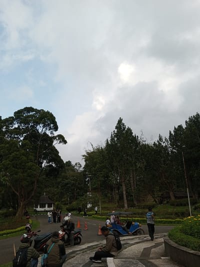 The image depicts an outdoor scene in Ambositra, Madagascar, under a sky with both blue patches and substantial grey cloud cover. In the foreground, a paved road curves uphill from left to right, bordered by a concrete path and green vegetation, including yellow flowers on the right. On the left side of the road, two individuals are crouched near a black scooter/motorcycle, with a second black scooter/motorcycle parked adjacent to them. A third male individual, wearing a light brown jacket and dark pants with a black backpack, is seated on a low concrete barrier on the right, looking at a mobile device.

In the midground, two orange traffic cones are placed on the paved road. A blue, three-wheeled motorized vehicle with a dark canopy is visible on the right, carrying multiple passengers. Further uphill, a person wearing a striped shirt and dark pants is walking away from the camera. A small, white building with a dark, ornate roof is situated on the left, nestled among trees. The background is characterized by dense green foliage and numerous trees, some tall and slender, covering hilly terrain. A utility pole is visible on the right side of the scene.