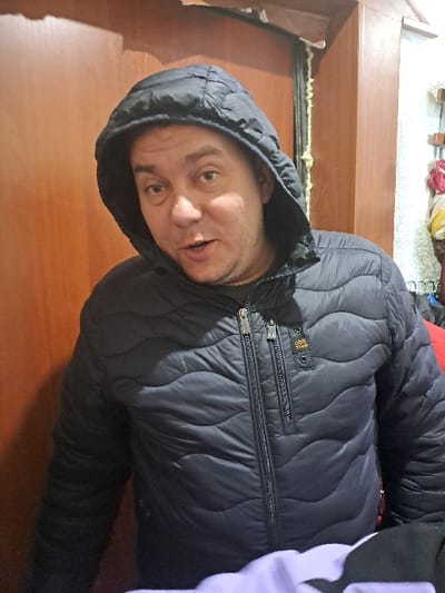 This image is located in Bolshaya Elhovka, Russia. It depicts a single adult male, approximately mid-frame, in an indoor setting. The male is Caucasian, with short dark hair partially obscured by a hood, light skin, and visible stubble on his face. His mouth is slightly open, and his eyes are directed forward, towards the camera. He is wearing a dark blue or black quilted puffer jacket with a hood, featuring a full-length central zipper and additional zippers on the chest. A small black label with light text, possibly "GSR SWO," is present on the upper right chest of the jacket. A grey garment is visible beneath the partially unzipped jacket. The background to the left consists of a light brown wooden panel or door. To the right, a wooden doorframe is visible, with exposed light-colored expanding foam sealant along its edge. Beyond the doorframe, a lighter-colored wall is present, displaying indistinct colorful fabrics (red, yellow, white) and a wooden fixture in the upper right corner. A segment of light purple fabric is visible in the lower right foreground.