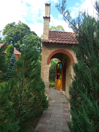 An outdoor, eye-level, full shot shows a path leading to an arched entryway of a brick building. The entryway has a wooden door with stained glass panels and an open archway above it, revealing more greenery beyond. The building has a tiled roof and a brick chimney. The path is paved with square tiles of gray and red. Lush green trees and bushes surround the building, with a clear blue sky with white clouds overhead. The scene is set in the city of Giaginskaya, Russia.