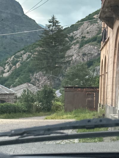 The image shows a rocky, forested mountainside in Tumanyan, Armenia. The scene is oriented vertically, suggesting it was taken while the camera was tilted or the image has been rotated. In the foreground, a grey, textured surface, possibly a road or path, runs alongside a grassy verge. A dark railing or fence runs along the right side of the frame, casting shadows on the ground.

Further back, the mountainside is covered in dense green trees, with visible rocky outcrops and slopes. A white object, possibly a kite or a piece of cloth, is suspended on a thin line stretched across the scene, adding a touch of dynamic activity to the otherwise static landscape. There's a small, rustic wooden building with a dark door visible amongst the trees. In the distance, the sky is overcast with clouds. The overall impression is a natural, somewhat rugged, outdoor setting. No people are visible in the frame. The lighting suggests it is daytime.