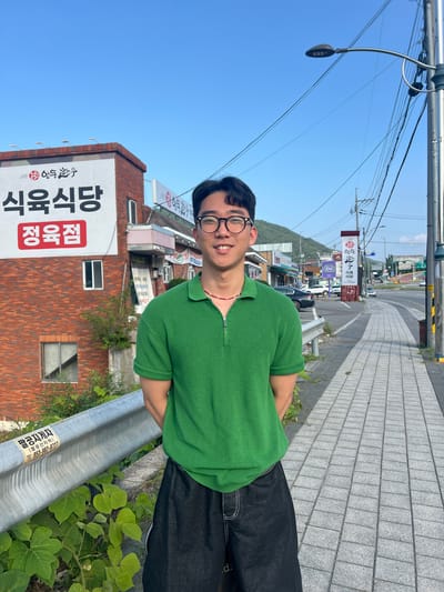 A young East Asian man wearing glasses, a green polo shirt, and black shorts stands on a sidewalk in front of a building. The building, which appears to be a restaurant, has a large sign with Korean text. The man is smiling and looking at the camera. The sidewalk is paved with gray tiles, and there is a metal guardrail to the left of the man. The background shows a street with cars and more buildings, as well as a blue sky and some distant hills. The location is identified as Dongmyeong-myeon, South Korea.