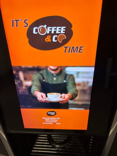 The image displays a digital advertisement for "Coffee & Co" time. The top portion of the advertisement features the text "IT'S" and "COFFEE & CO TIME" in large, stylized font. A logo for "Coffee & Co" is visible, incorporating a coffee cup and a bite mark on a cookie. Below this, a video feed shows a barista in a green shirt and dark apron holding a white cup of coffee with latte art on a saucer. The bottom section of the advertisement provides contact information for "Coffee & Co" in Mechelen, Belgium, including their address at Blarenberglaan 23A, postal code 2800, phone number 015 32 30 03, and email address info@coffeeandco.be. The image is framed by the dark exterior of a digital display or machine. The location in the city of Boom, Belgium is not explicitly mentioned in the image itself.