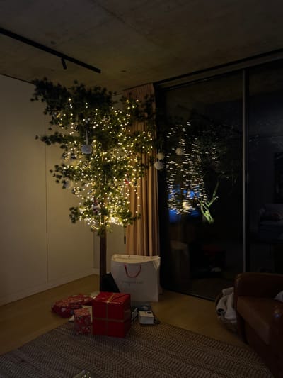 A decorated Christmas tree stands next to a large window, reflecting exterior lights. Gifts, wrapped in red and patterned paper, are scattered around the base of the tree, alongside a white shopping bag. A rug covers part of the floor and a brown chair sits in the background. The scene is illuminated by the warm glow of the tree lights.